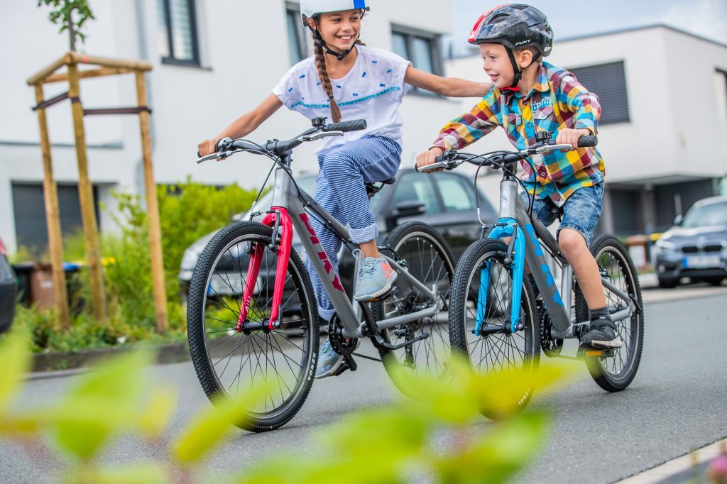 Ein Mädchen und ein kleinerer Junge fahren auf Fahrrädern. Das Mädchen lächelt dem Jungen zu und hat eine Hand auf seiner Schulter, um ihn zu unterstützen.