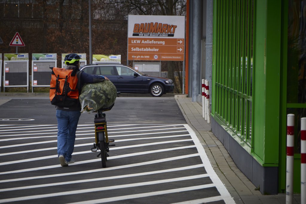 Eine Person schiebt ein Fahrrad mit einem verpackten Weihnachtsbaum vorm Eingangsbereich eines Baumarktes entlang.