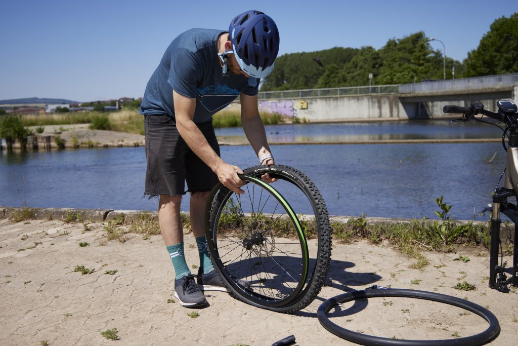 Ein Radfahrer steht vor einem Gewässer und zieht einen Fahrradreifen auf ein Vorderrad. Am Boden daneben liegt ein leicht aufgepumpter Fahrradschlauch.