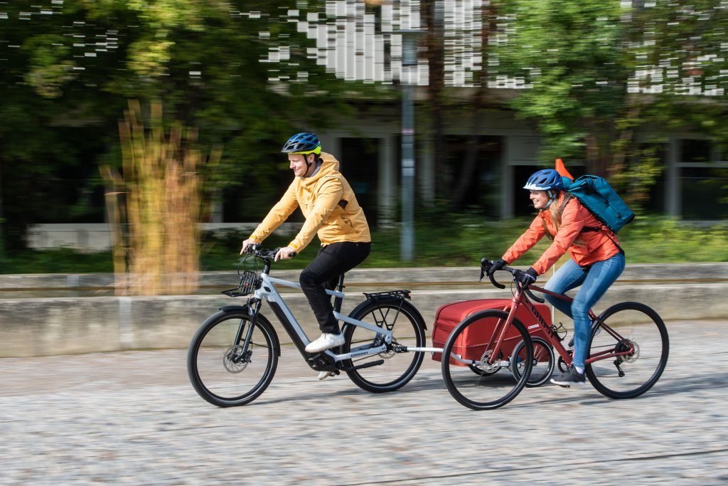 Ein Mann auf einem E-Bike mit Anhänger und eine Frau auf einem Rennrad fahren nebeneinander einen gepflasterten Weg entlang.