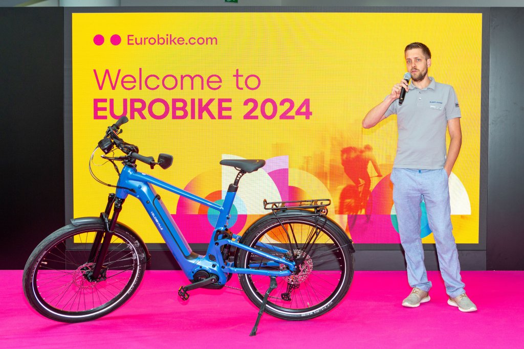 Mann auf einer Bühne auf der Eurobike mit Mikrofon vor einem E-Bike.