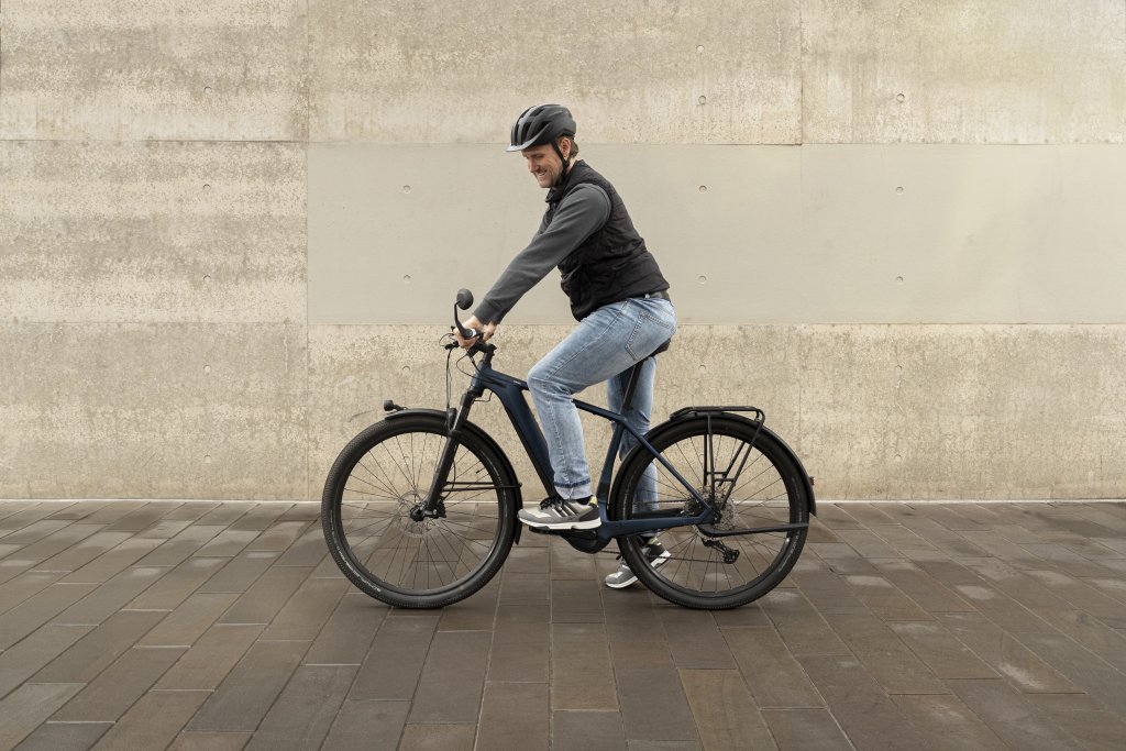 Ein Mann steht mit einem E-Bike neben einer Betonwand und schaut in den Rückspiegel am Lenkerende.