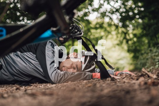 Eine Person mit Fahrradhelm liegt im Wald auf dem Boden. Im Vordergrund sind Teile eines Fahrrads zu erkennen.